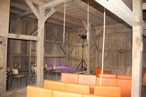 Die lila beleuchtete Bühne im Hintergrund, davor orange Sofas. Von oben hängen Seile, im Hintergrund steht eine Leuchte.