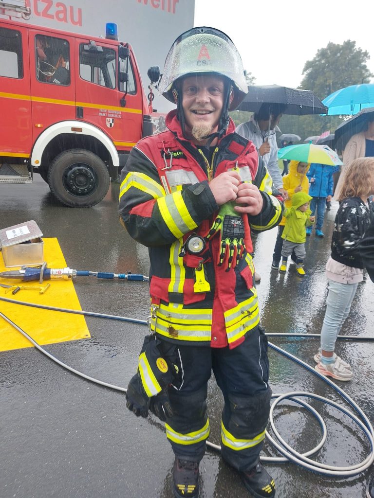 Das Foto zeigt den ehrenamtlichen Feuerwehrmann Domenik Kügler von der Feuerwehr Oberkotzau in seiner Ausrüstung. Links hinter ihm sieht man einen Ausschnitt eines Feuerwehrautos