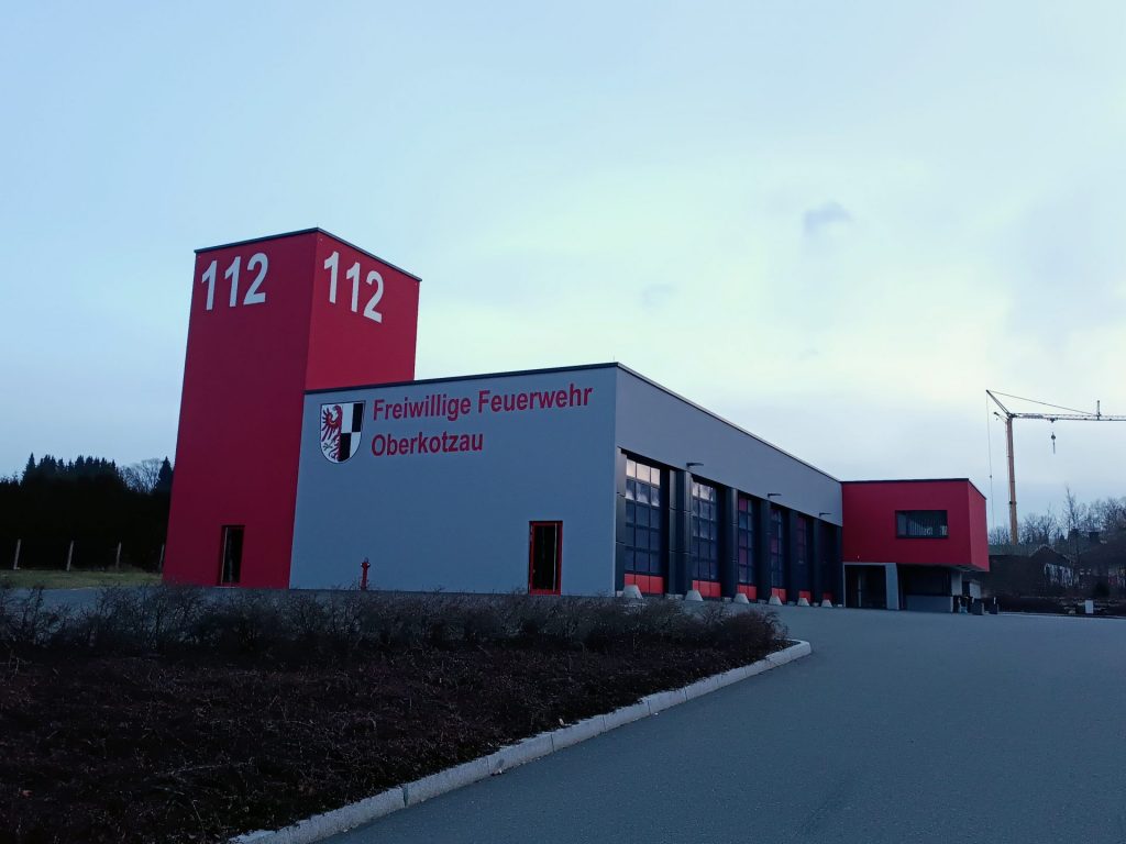 Das Foto zeigt das Gebäude der Freiwilligen Feuerwehr Oberkotzau. Mittig sieht man mehrere Tore, links überstrahlt der rote Schlauchturm mit der Notrufnummer 112