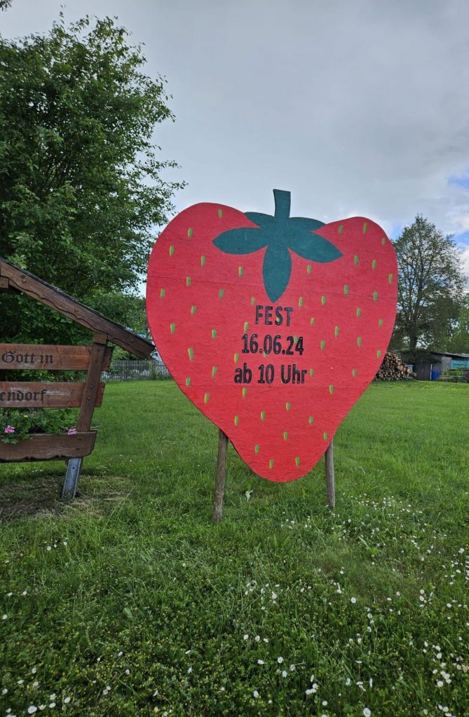 Das Foto zeigt links im Bild das Begrüßungsschild von Kautendorf am Ortseingang. Daneben sieht man eine große rote Erdbeere, in der der aktuelle Termin des Erdbeerfestes steht.
