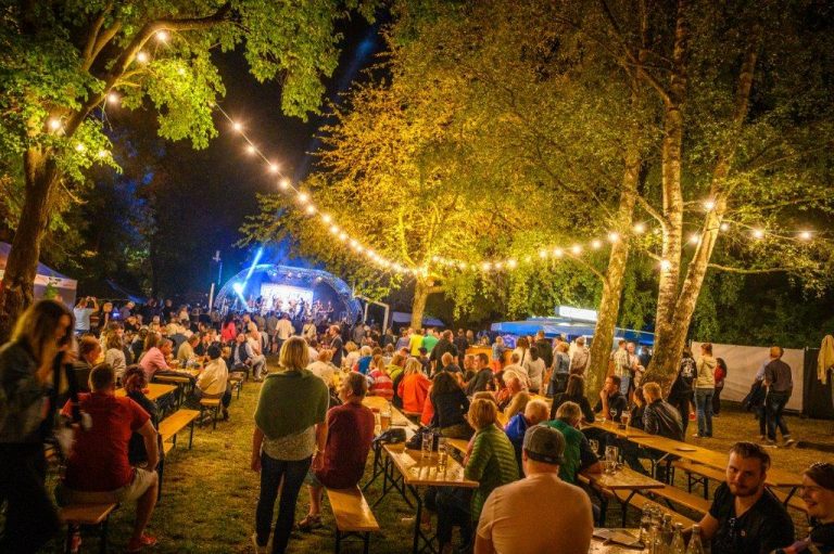 Das Foto zeigt das Saaleauenfest mit vielen Bierbänken und Besuchern. Lichterketten sind zwischen Bäumen aufgehängt. Im Hintergrund steht eine Bühne. Es ist schon dunkel und man spürt die Stimmung durch das Foto.