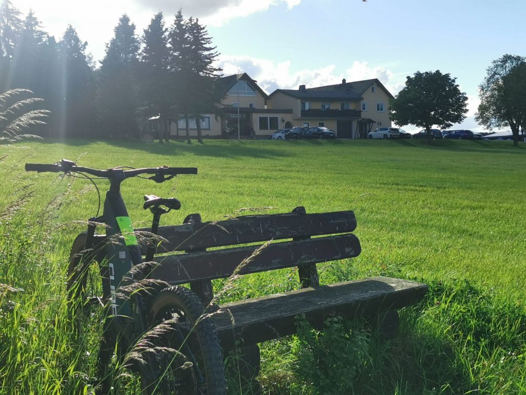 Einkehrmöglichkeit bei der Landkreisumrundung: Fahrrad lehnt an einer Holzbank. Im Hintergrund sieht man einen Gasthof hinter einer Wiese