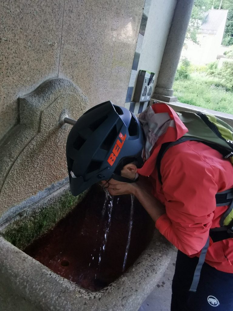 Frau mit Fahrradhelm trinkt aus einer Quelle mit eisenhaltigem Wasser