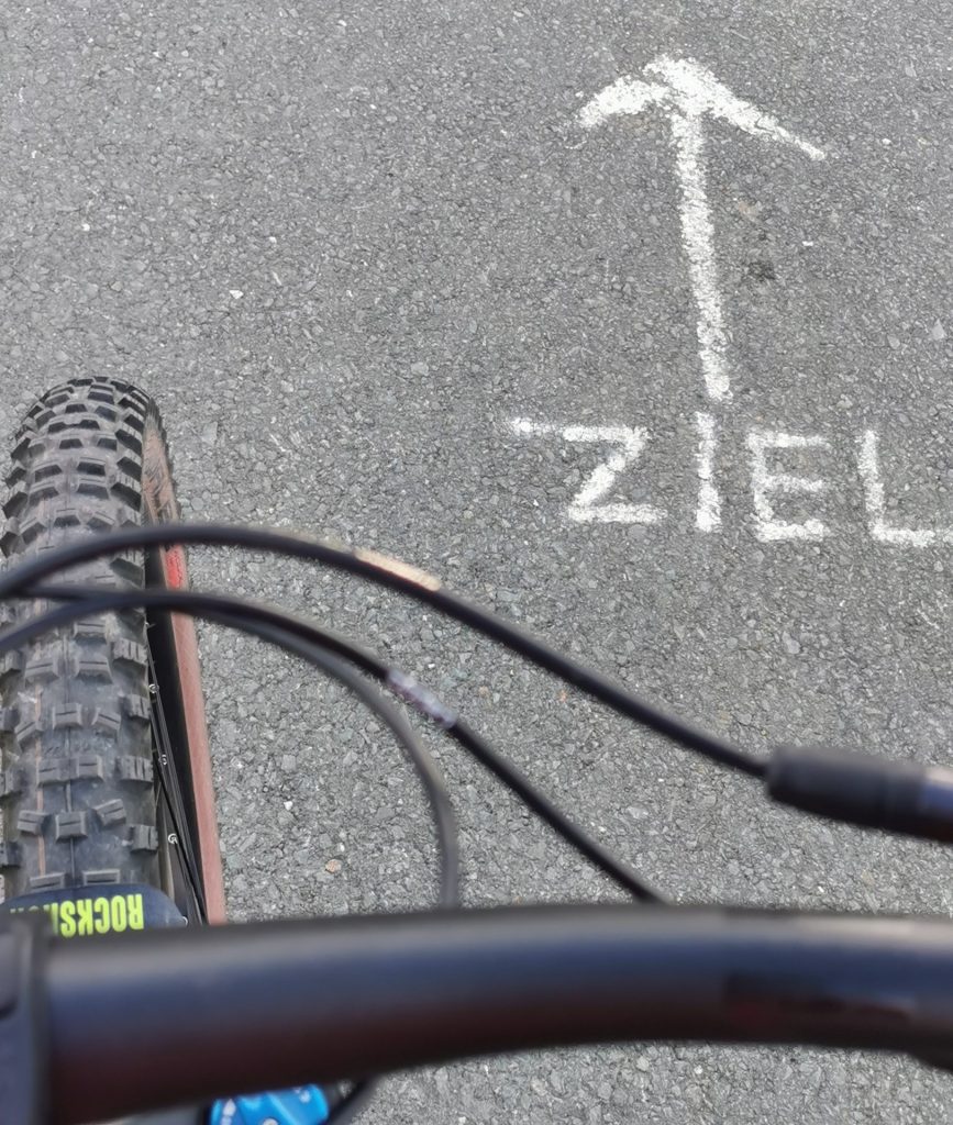 Vorderrad eines Fahrrads auf einer Straße, auf der mit Kreide "Ziel" geschrieben wurde