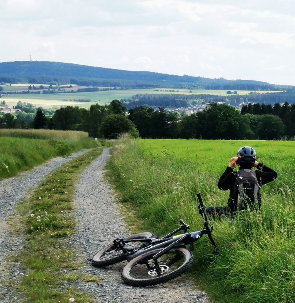 Fahrradfahrer fotografiert den Ausblick auf die Landschaft. Sein Fahhrad liegt daneben auf einem Feldweg.