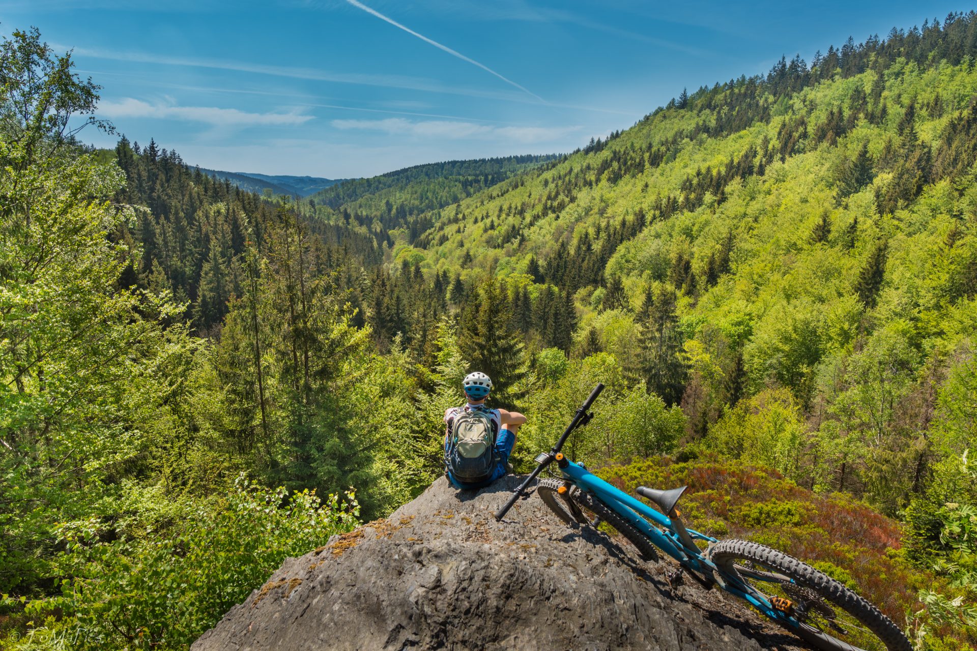 Bild zweigt Landschaft mit Fahrrad darauf und Fahrradfahrer sitzt davor auf einem Felsen und schaut in en grünes Tal mit Bäumen