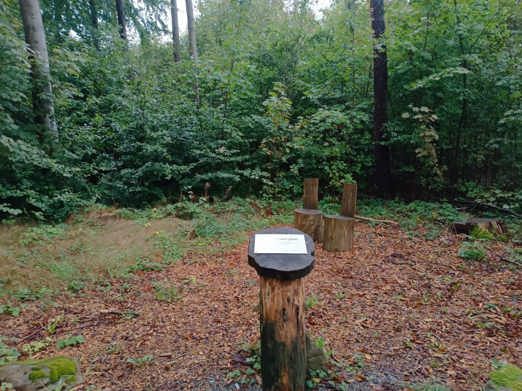 Das Foto zwei kleine Stühle, die aus Holzstümpfen gemacht sind. Vorne ist ein Holzstumpf mit dem Schild zum Neubeginn.