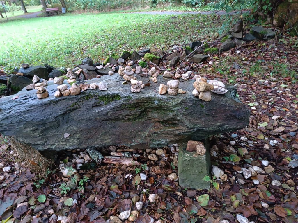 Dominant im Foto ist ein großer Stein, der als Tisch genutzt wird und auf dem Spaziergänger kleine Türmchen aus Steinen gebaut haben.