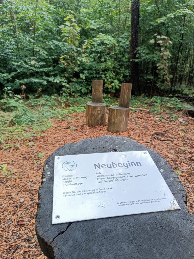 Das Foto zeigt ein Schild am Energieplatz "Neubeginn“, befestigt auf einem Holzstumpf. Das Schild erklärt die Wirkung des Elements Erde, das mit diesem Platz verbunden ist. Es beschreibt die stabilisierende und aufbauende Energie des Ortes, die Gefühle wie Friede, Geborgenheit, Ruhe und Heiterkeit hervorrufen kann.