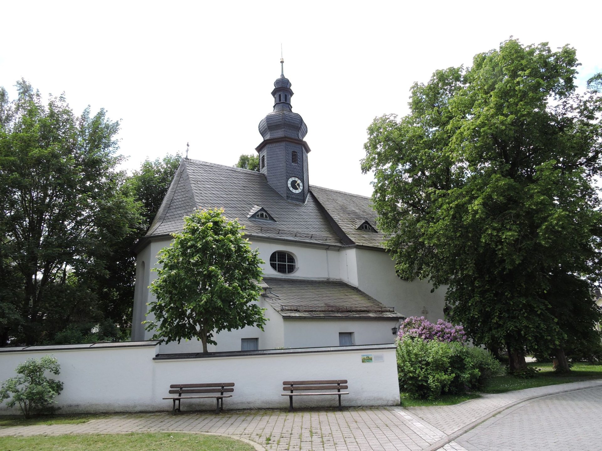 Auf dem Bild ist eine weiße Kirche mit Kirchturm zu sehen. Im Vordergrund sind außerdem zwei Laubbäume mit grünen Blättern.